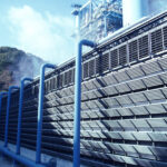 Blue-painted industrial facility with a row of vertical pipes and a slatted metal facade, steam rising near the hills in the background.