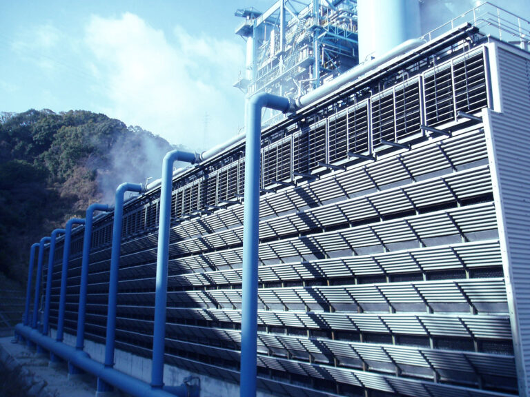 Blue-painted industrial facility with a row of vertical pipes and a slatted metal facade, steam rising near the hills in the background.
